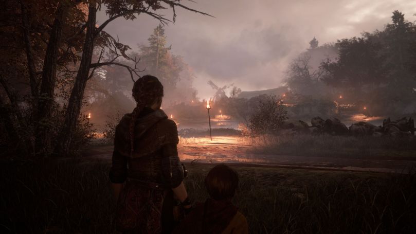Amicia and Hugo trying to reach a windmill in the far distance. The field is covered by torches lighting the way at dusk.